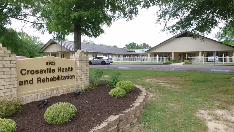 Exterior view of Crossville Health and Rehabilitation facility showing a beige brick sign with the facility name and address 8922, a parking lot with a parked car, and single-story buildings with a white fence and trees in the background.