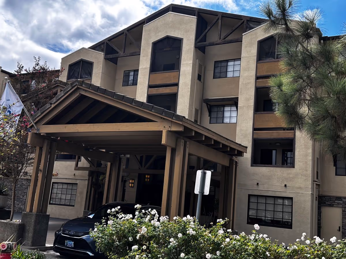 Entrance canopy and facade of a multi-story senior living building with a parked car and flowering landscaping in front.