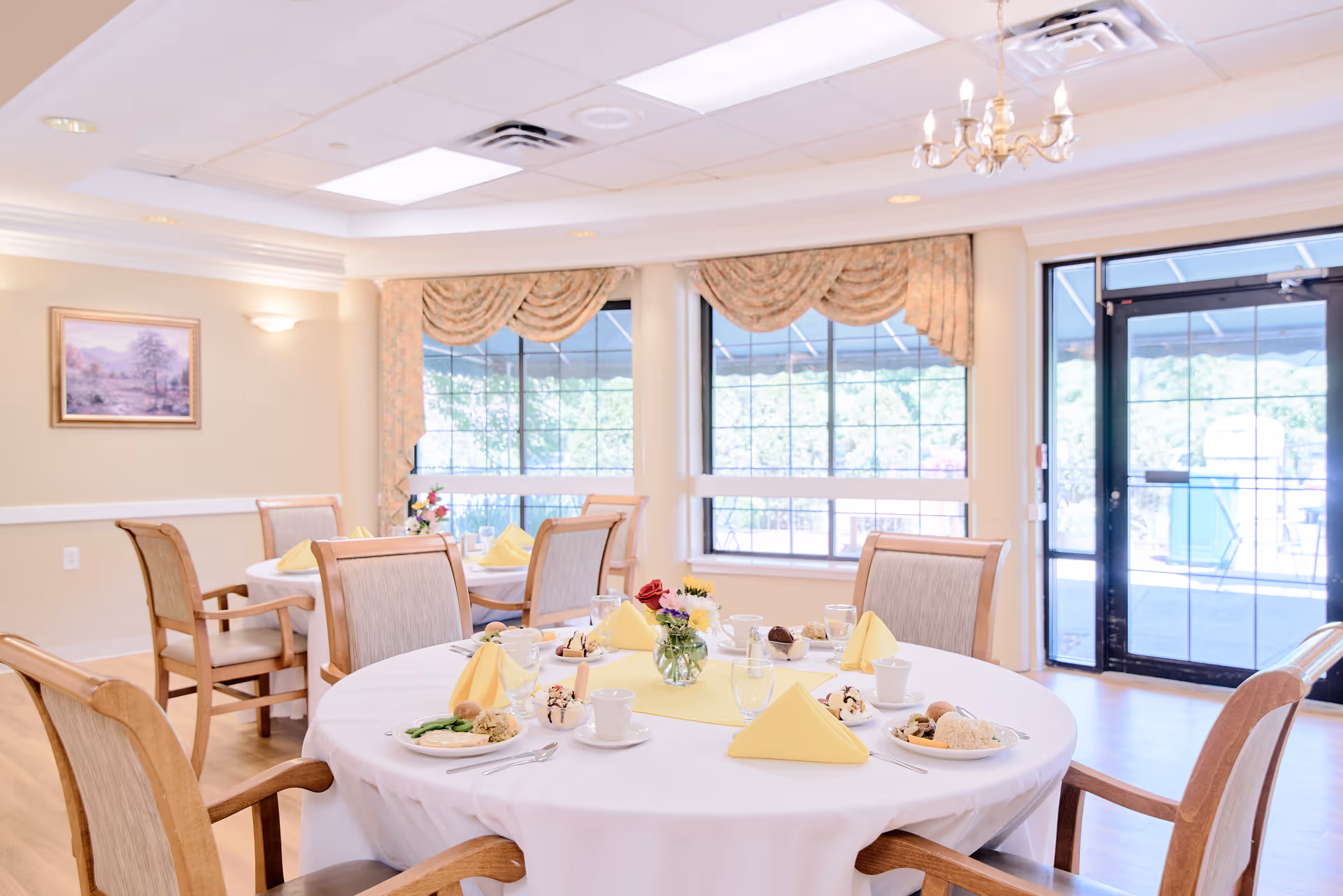 A bright dining room with round tables covered in white tablecloths, set with plates of food, cups, glasses, and yellow folded napkins. The room has large windows with floral curtains, wooden chairs with cushioned seats, a chandelier, and a glass door leading outside.
