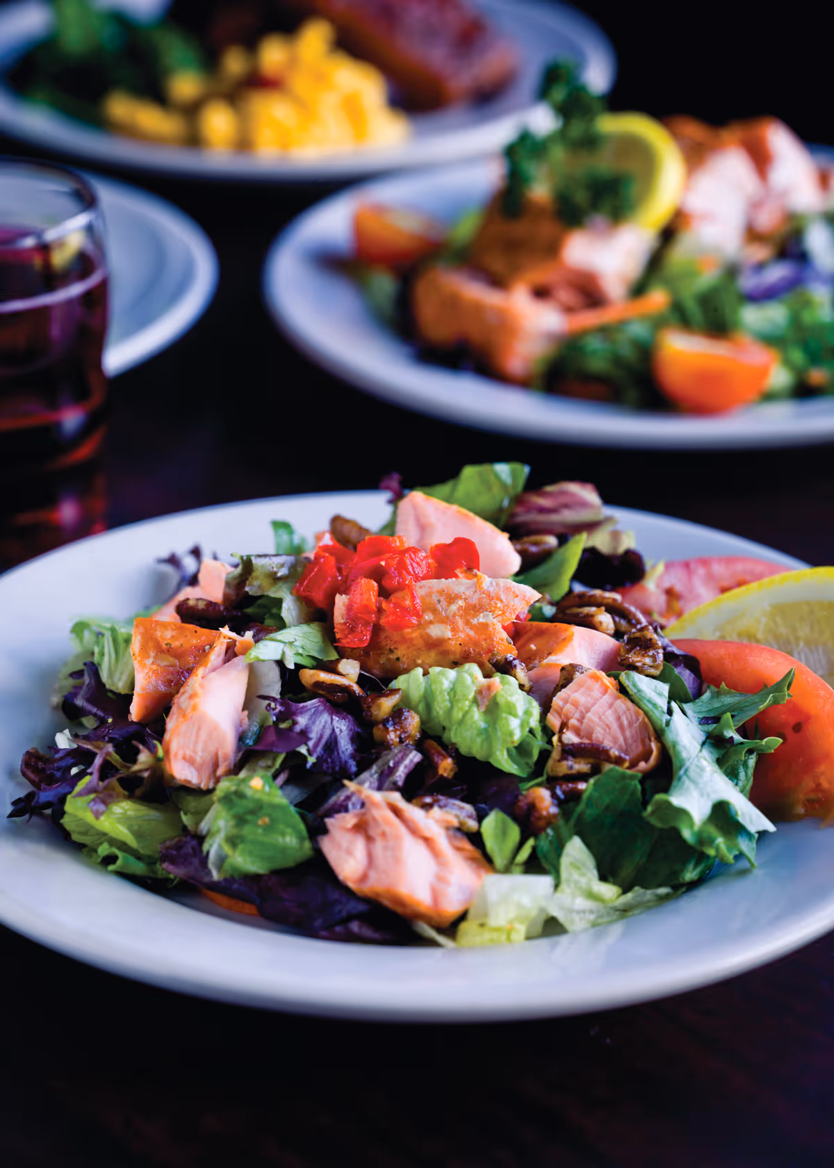 A close-up of a fresh salad with mixed greens, pieces of cooked salmon, chopped red bell peppers, and pecans on a white plate, with a lemon wedge and tomato slice on the side. In the background, there are blurred plates of food and a glass of beverage on a dark wooden table.