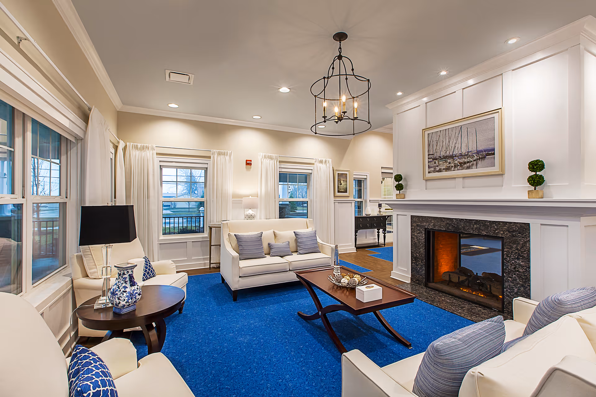 A cozy living room with cream-colored sofas and armchairs, blue cushions, a blue carpet, a wooden coffee table, and a fireplace with a framed picture above it. Large windows with white curtains allow natural light to enter the room.