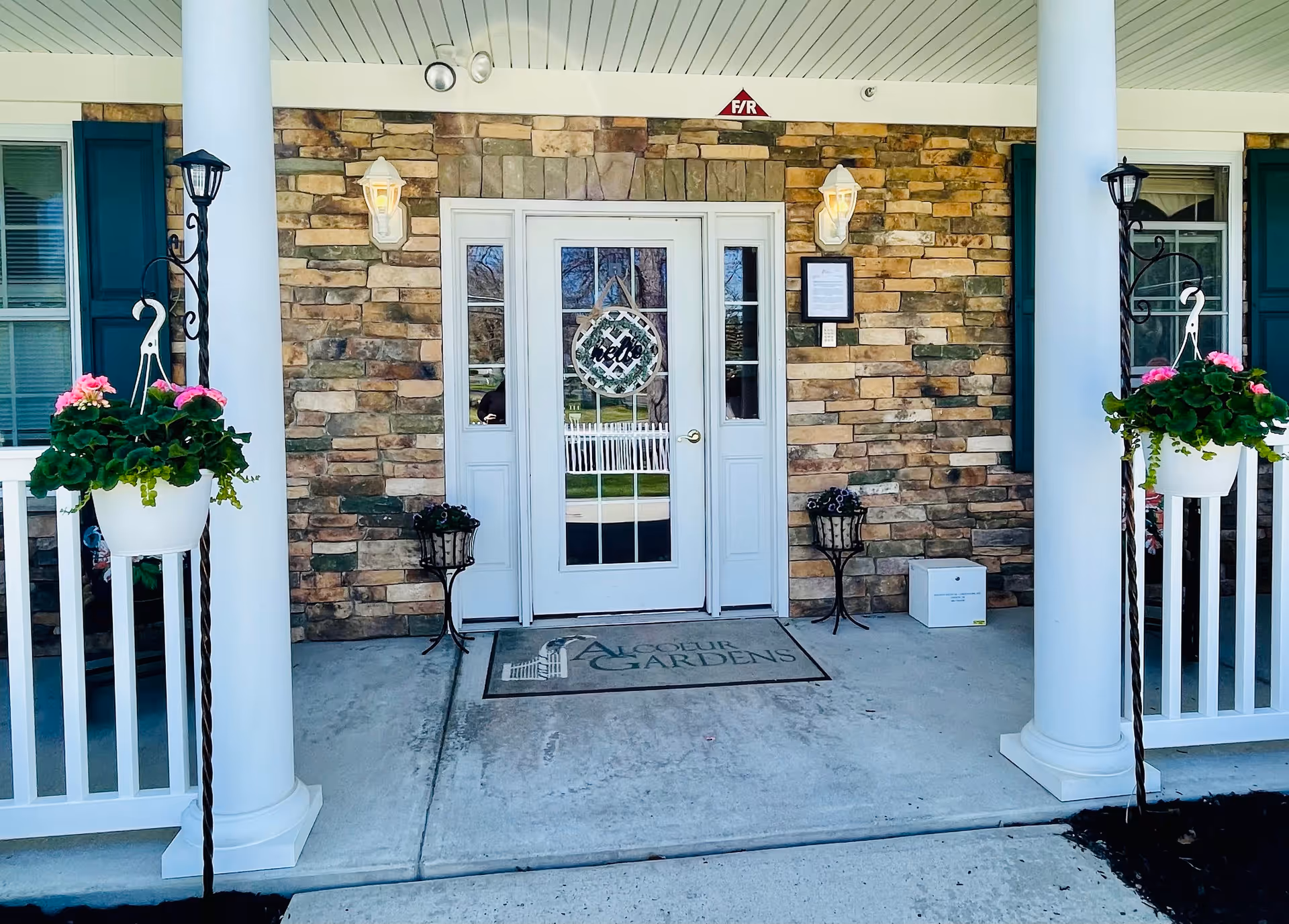 Entrance to Alcoeur Gardens at Toms River featuring a white door with glass panels, stone wall exterior, two white columns, hanging flower pots with pink flowers, two wall-mounted lantern lights, and a welcome mat with the facility's name.