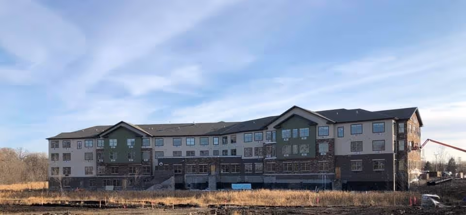 Wide exterior view of a three-story building under construction with beige and green walls, multiple windows, and a dark roof, surrounded by dry grass and construction equipment under a partly cloudy sky.