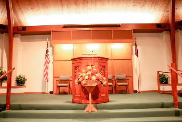 Interior chapel sanctuary showing a wooden pulpit with a floral arrangement on a raised platform flanked by chairs and flags.
