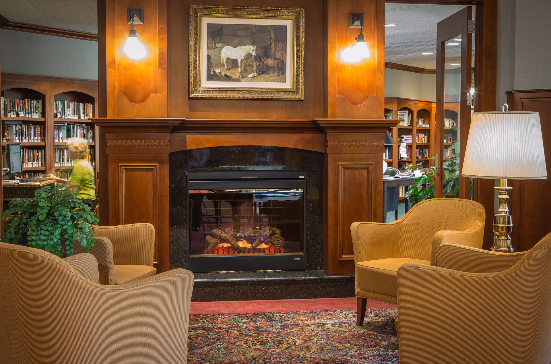Cozy senior living community lounge area with four beige armchairs arranged around an electric fireplace set in a wooden mantel. Above the fireplace is a framed painting of horses. The room features warm lighting from wall sconces and a table lamp. In the background, a woman is browsing bookshelves in a library area.