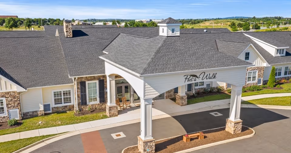 Front exterior view of Poet's Walk Leesburg, A Memory Care Community, showing a covered entrance with white pillars and stone bases, a paved driveway, and a building with stone and white siding under a gray shingled roof.