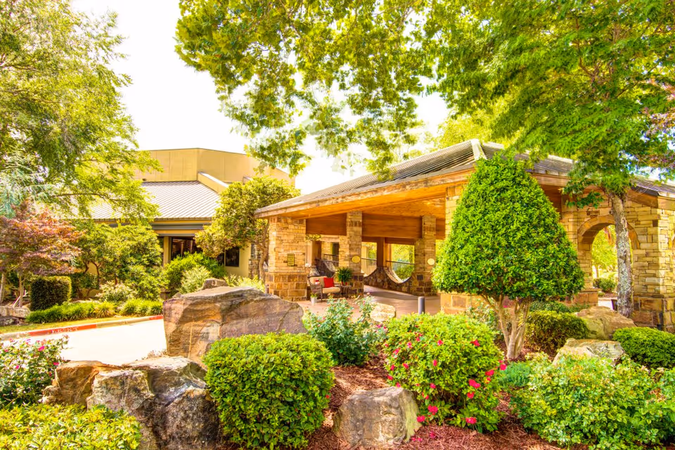 Exterior view of The Atrium at Serenity Pointe featuring a covered entrance with stone pillars and wooden roof, surrounded by lush green trees, bushes, flowering plants, and large rocks in a landscaped garden.