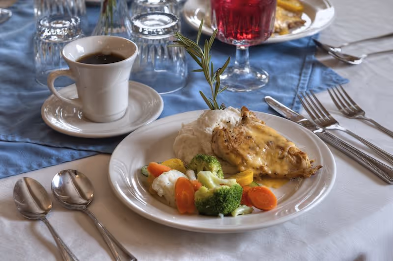 A plated meal consisting of grilled chicken breast with melted cheese, mashed potatoes, and steamed mixed vegetables including broccoli, carrots, and cauliflower, served on a white plate. Next to the plate is a cup of black coffee on a saucer, with silverware and glasses on a table covered with a white tablecloth and a blue cloth underneath some of the items.