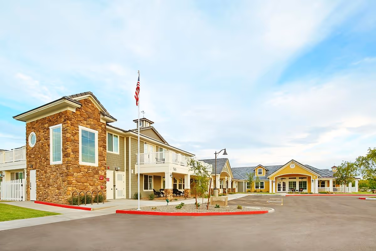 Exterior view of American Orchards Assisted Living & Memory Care facility showing a two-story building with stone and siding facade, an American flag on a flagpole, a parking area, and a clear sky with some clouds.