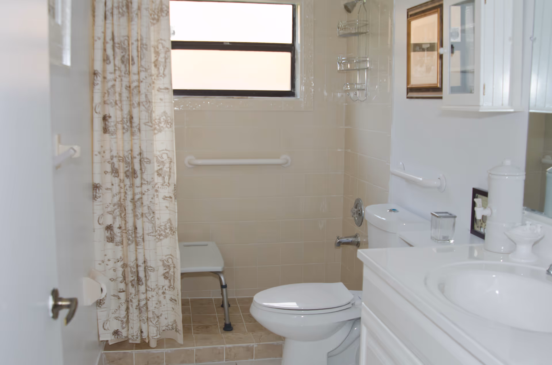 A small bathroom with a white toilet, a white sink with a countertop, a shower area with a beige tiled wall, a shower curtain with a map design, a shower chair, and grab bars on the walls for support. There is a frosted window above the shower area and a framed picture on the wall.