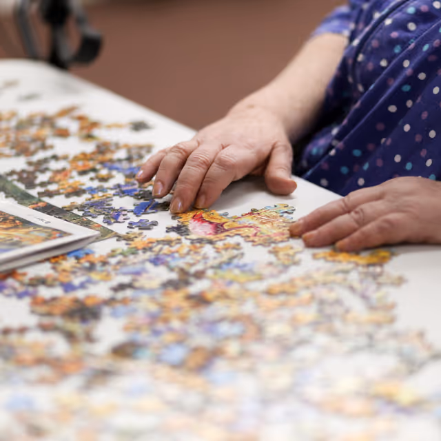 Close-up of an elderly person's hands assembling a colorful jigsaw puzzle on a white table, with scattered puzzle pieces and part of the puzzle box visible.