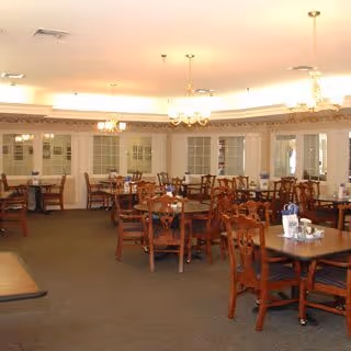 Spacious dining room with multiple wooden tables and chairs, place settings, chandeliers, and large windows along the back wall.