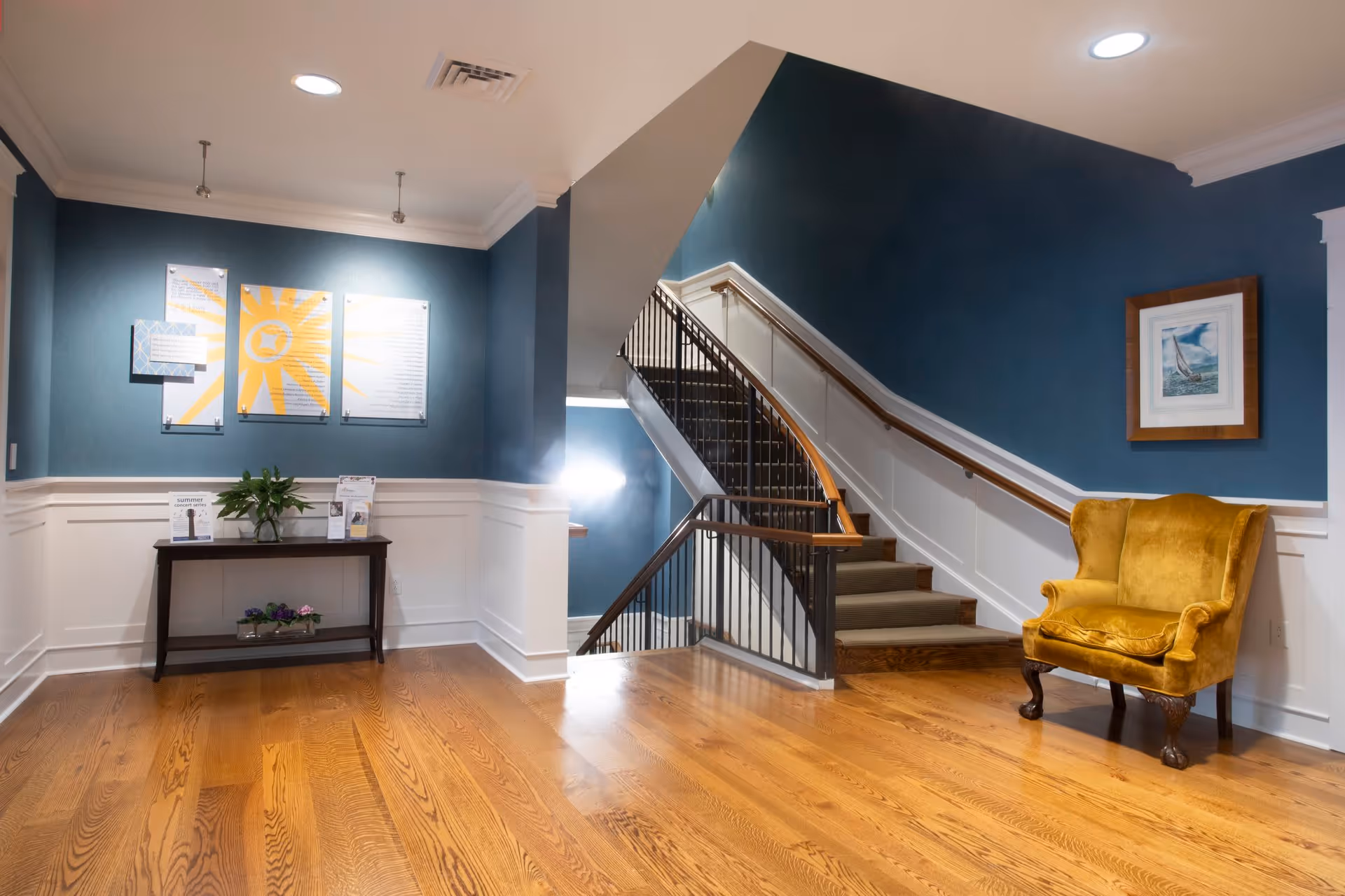 Interior view of a senior living facility with wooden flooring, blue walls with white wainscoting, a staircase with a wooden handrail and black balusters, a yellow upholstered armchair, a framed picture on the wall, and a small table with plants and informational materials under three decorative wall panels.