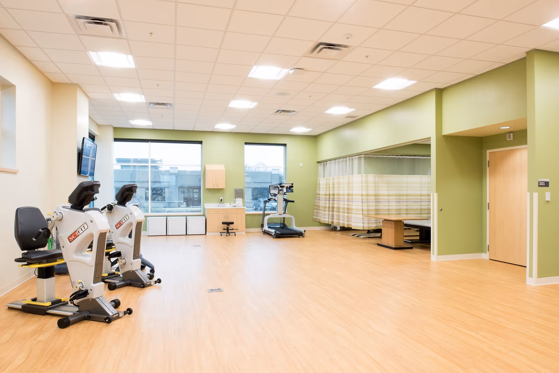 A spacious rehabilitation room with light wood flooring and green walls. The room contains two stationary exercise bikes, a treadmill, a small desk with a stool, and a curtained area with a treatment table. Large windows let in natural light, and a wall-mounted TV is visible on the left side.