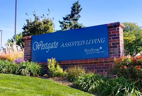 Blue Westgate Assisted Living sign on brick wall with landscaping and flowers.