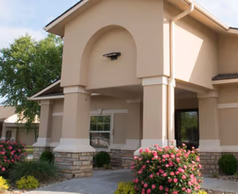 Exterior view of a beige building entrance with a covered porch supported by columns. There are bushes with pink flowers and green shrubs near the walkway leading to the entrance. The sky is partly cloudy.
