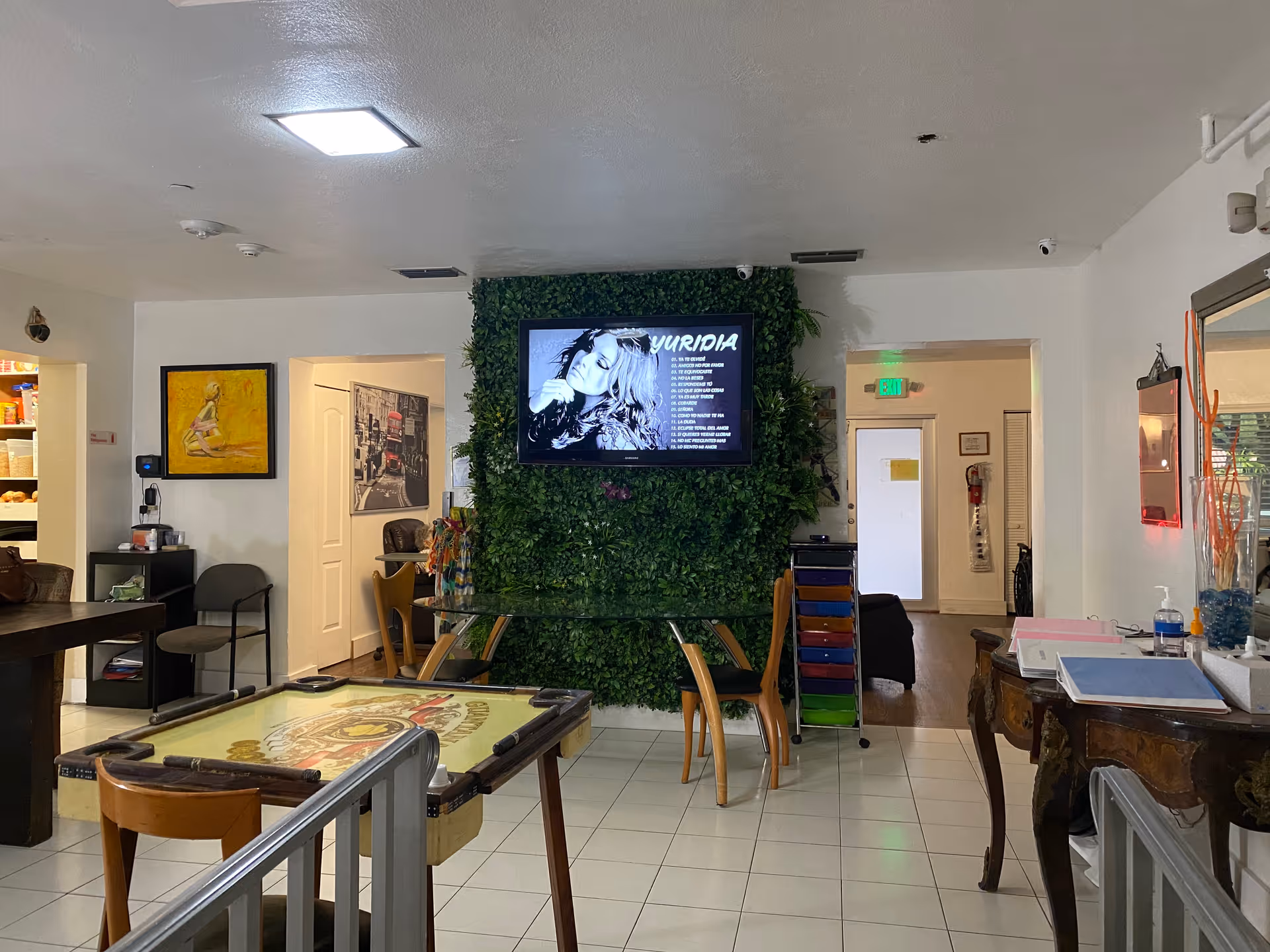 Interior view of a common area in a senior living facility with a pool table in the foreground, a glass table with wooden chairs, a wall covered with green foliage and a mounted TV displaying a music playlist. The room has white tiled floors, various pieces of furniture including chairs and a wooden desk with papers and a hand sanitizer bottle, and artwork on the walls.