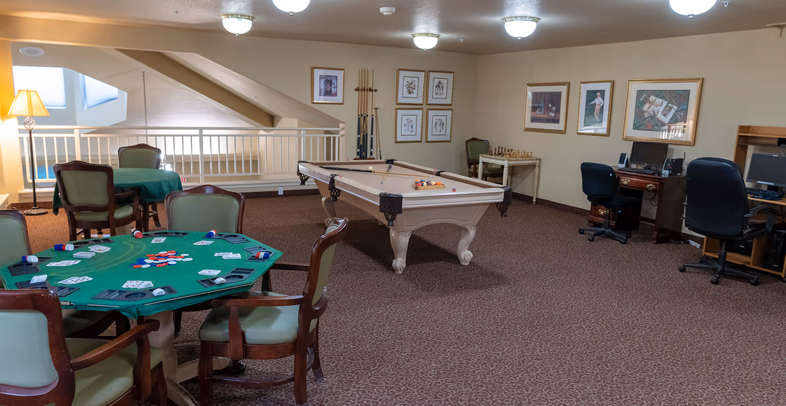 Carpeted community game room with a poker table, a pool table, chairs, and computer desks in a senior living facility.