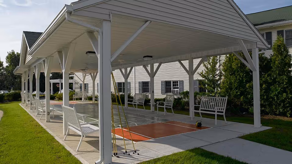 Covered outdoor shuffleboard court with white benches along the sides, adjacent to a white building with windows and surrounded by green grass and shrubs.