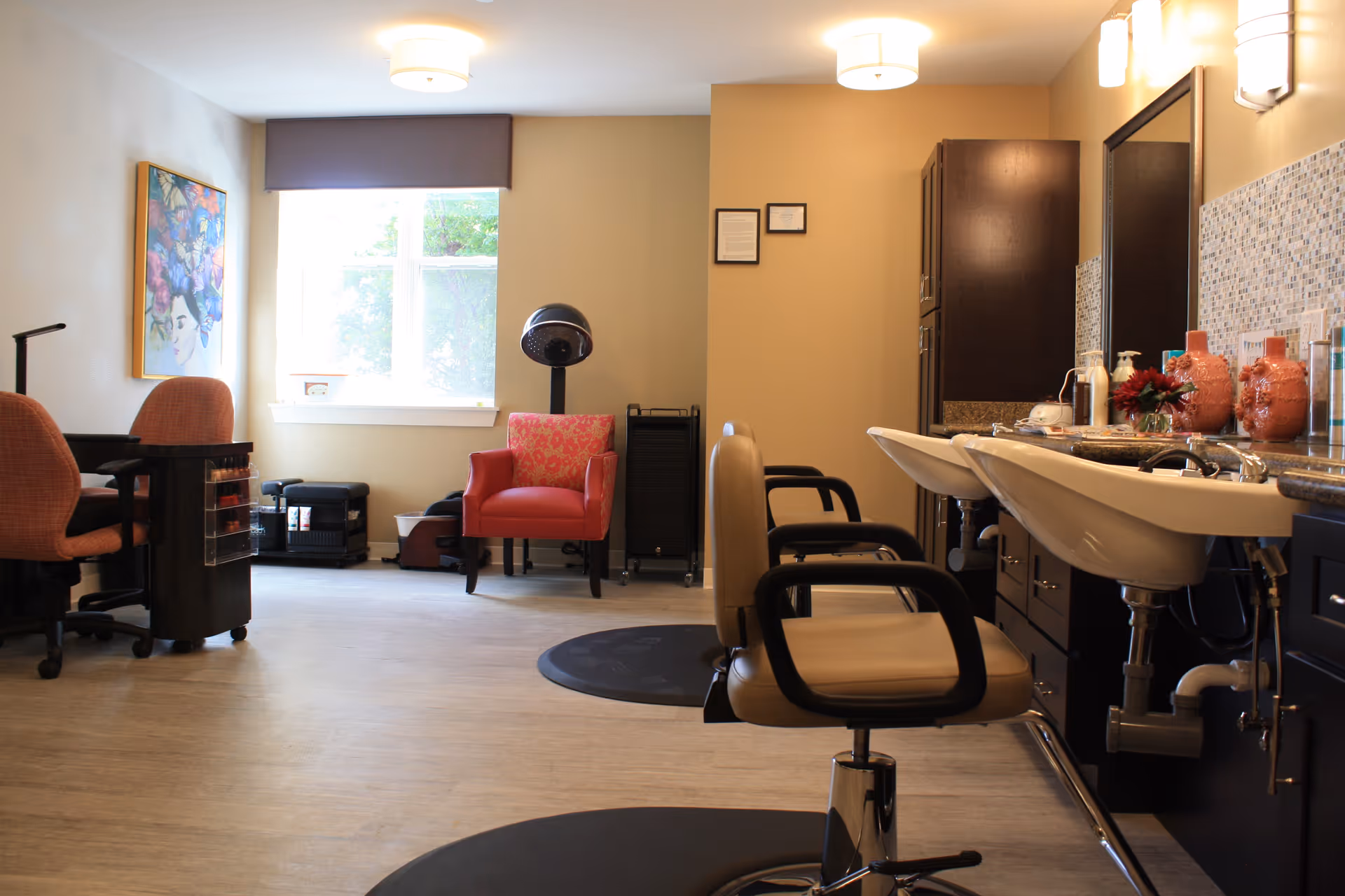 Interior view of a salon area in a senior living facility with two salon chairs in front of sinks, a red armchair with a pink cushion near a window, a hair dryer, and a nail station with two orange chairs and a colorful painting on the wall.