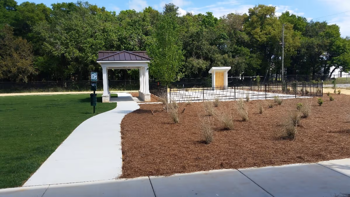 Outdoor area with a curved concrete walkway leading to a small white pavilion with a dark roof. To the right of the walkway is a mulched garden area with small shrubs and a fenced section containing a white structure. Trees and greenery are visible in the background under a partly cloudy sky.