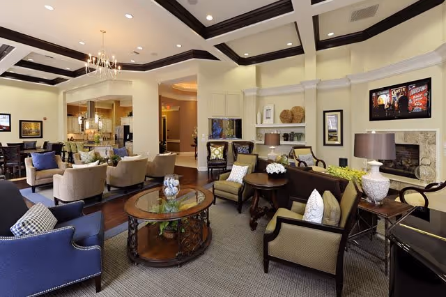 A spacious and elegantly furnished living room area with multiple armchairs and sofas arranged around glass-top coffee tables. The room features a high ceiling with dark wooden beams, a chandelier, built-in shelves with decorative items, a fireplace, and warm lighting. The background shows a kitchen area with pendant lights and a dining table with chairs.