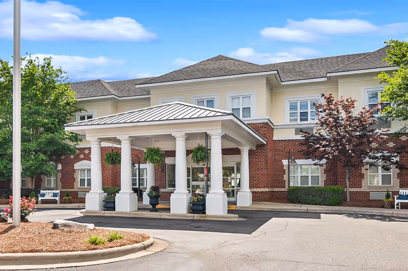 Exterior view of Morningside of Gastonia senior living facility showing a covered entrance with white columns, hanging plants, and a brick and beige building facade under a blue sky with some clouds.