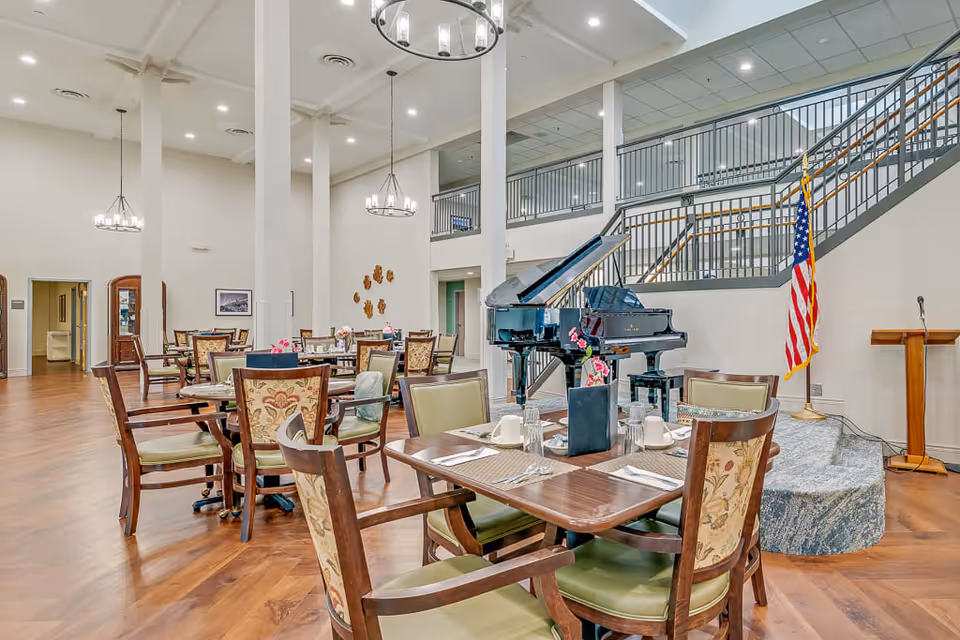 A spacious dining area in a senior living facility with multiple wooden tables and chairs arranged neatly. The room features high ceilings with hanging chandeliers, a grand piano on a small stage, an American flag, and a staircase with metal railings leading to an upper level. The floor is wooden, and the walls are painted white, creating a bright and welcoming atmosphere.