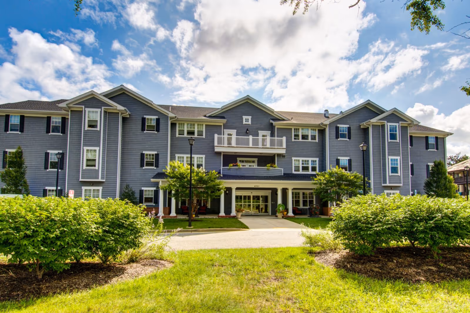 Front exterior view of a large, three-story senior living facility building with gray siding, white trim, and black shutters. The entrance is covered with a porch supported by white columns, and there are bushes and green grass in the foreground under a partly cloudy blue sky.
