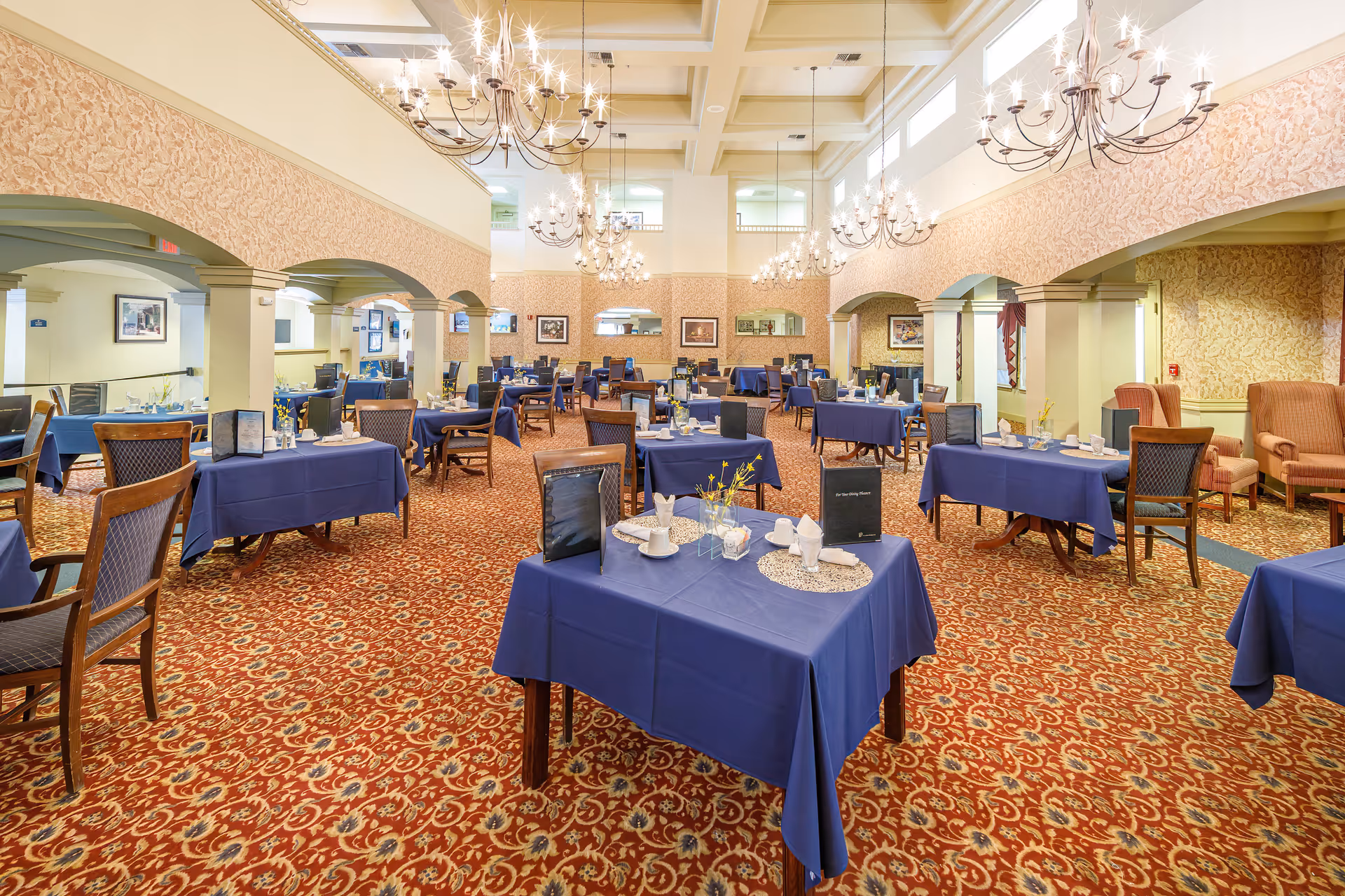 Spacious formal dining room with multiple tables set with blue tablecloths, chandeliers, and a patterned carpet.