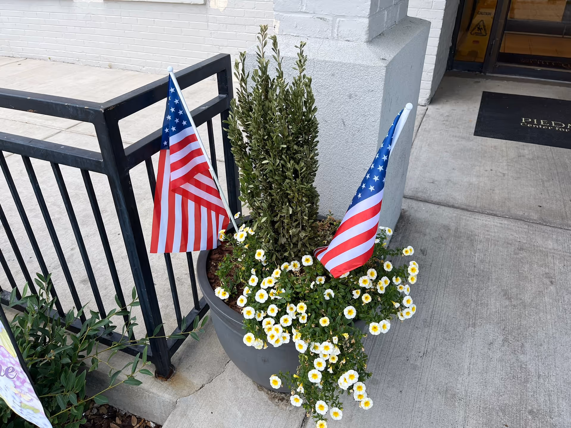 A large gray planter with green plants and white flowers with yellow centers, decorated with two small American flags, placed on a concrete surface near a black metal railing and a white brick wall. Part of a building entrance with a black mat is visible in the background.
