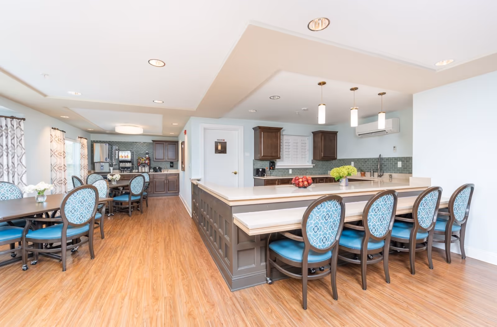 A bright and spacious dining area with wooden flooring, featuring a large kitchen island with a beige countertop and six blue cushioned chairs with patterned backs. The kitchen area has dark wooden cabinets, a green tiled backsplash, and modern pendant lights hanging from the ceiling. In the background, there are additional dining tables with matching chairs and windows with patterned curtains allowing natural light to enter.