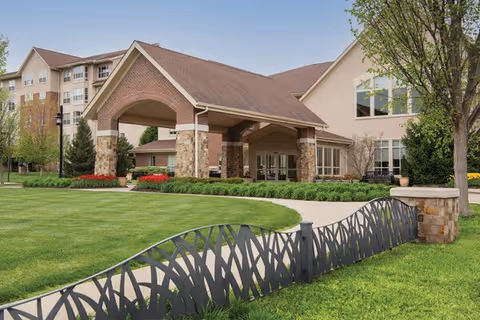 Exterior view of Tallgrass Creek Senior Living Community featuring a large covered entrance with brick and stone pillars, manicured green lawn, decorative metal fence, and trees under a clear sky.