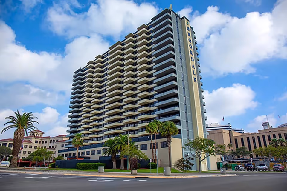 Tall multi-story residential building with balconies on each floor, surrounded by palm trees and other buildings under a partly cloudy blue sky.