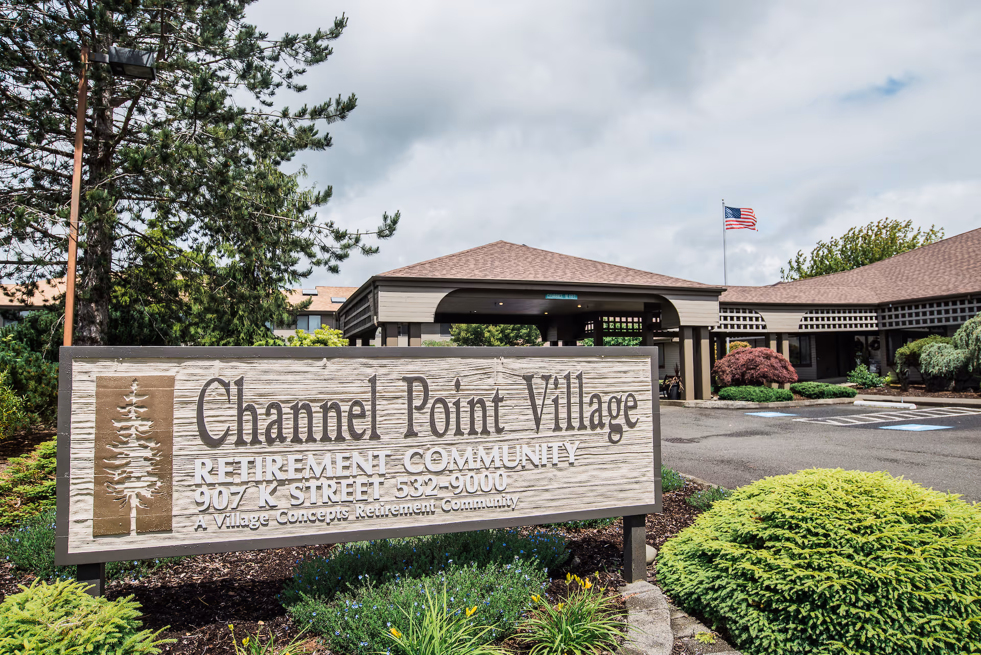 Entrance sign for Channel Point Village Retirement Community with the building and an American flag in the background, surrounded by greenery and landscaping.