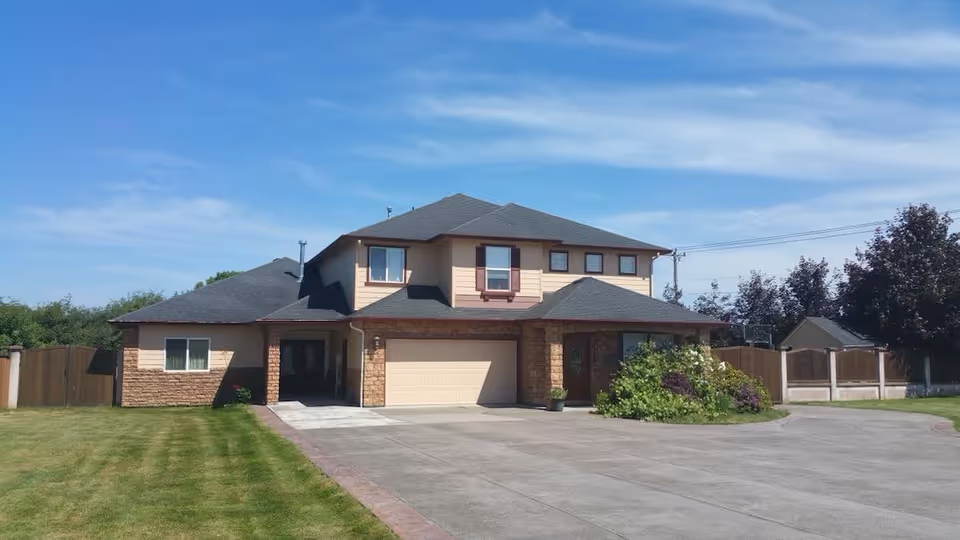 Two-story residential building with a large driveway, well-maintained lawn, and some bushes near the entrance under a clear blue sky.