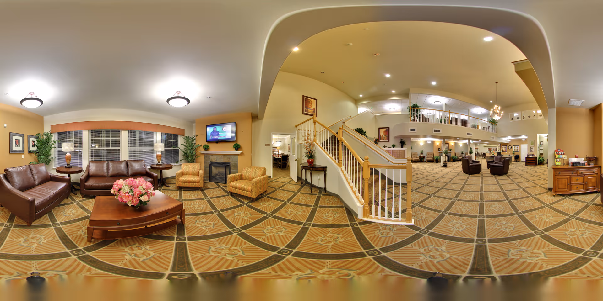 A spacious and well-lit senior living facility common area with a seating arrangement including leather sofas and armchairs around a coffee table with a flower arrangement. There is a fireplace with a TV mounted above it, large windows with blinds, and decorative plants. The area extends to a staircase leading to an upper level with railings, and additional seating areas are visible in the background. The carpet has a patterned design, and the walls are painted in warm tones.