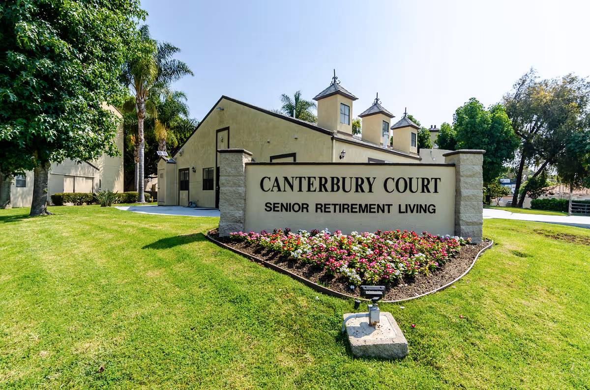 Exterior view of Canterbury Court Senior Retirement Living facility with a large sign in front surrounded by a flower bed and green lawn, trees and a beige building with three small tower-like structures in the background under a clear blue sky.