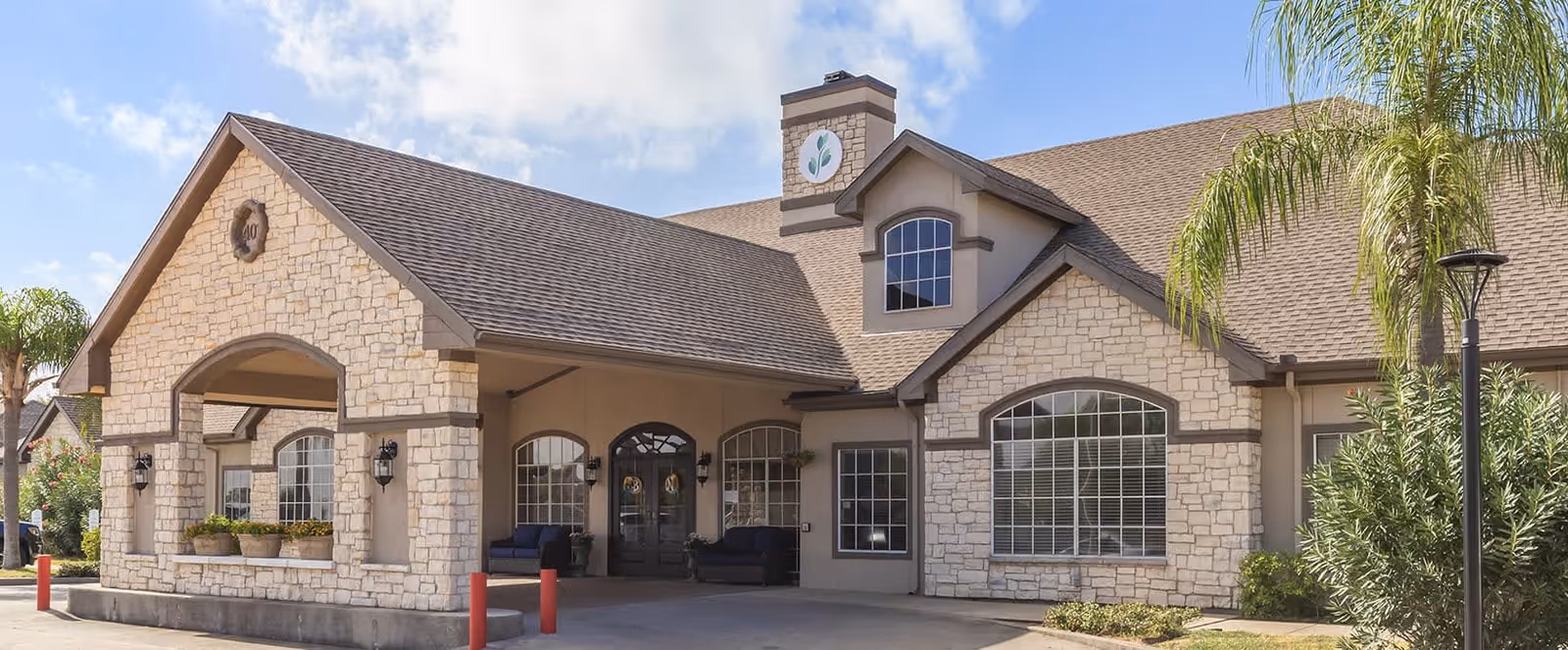 Exterior view of a senior living facility building with stone and stucco facade, large windows, and a covered entrance area. There are palm trees and landscaping around the building under a partly cloudy sky.