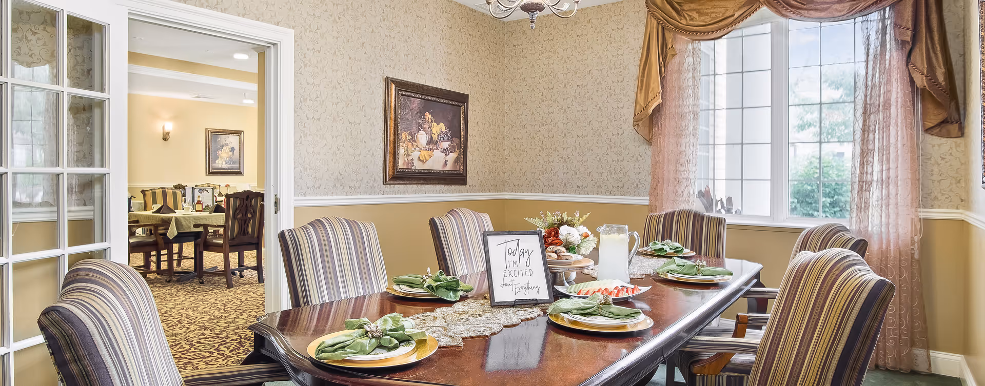 A formal dining room with a polished wooden table set for six people. Each place setting has a gold charger plate, a white plate, and a green cloth napkin with a decorative napkin ring. The table also features a lace table runner, a pitcher of lemonade, a plate of sliced strawberries, and a small floral centerpiece. A framed sign on the table reads 'Today I'm excited about everything.' The room has patterned wallpaper, a large window with sheer pink curtains, and a chandelier overhead. Through an open doorway, another dining area with tables and chairs is visible.