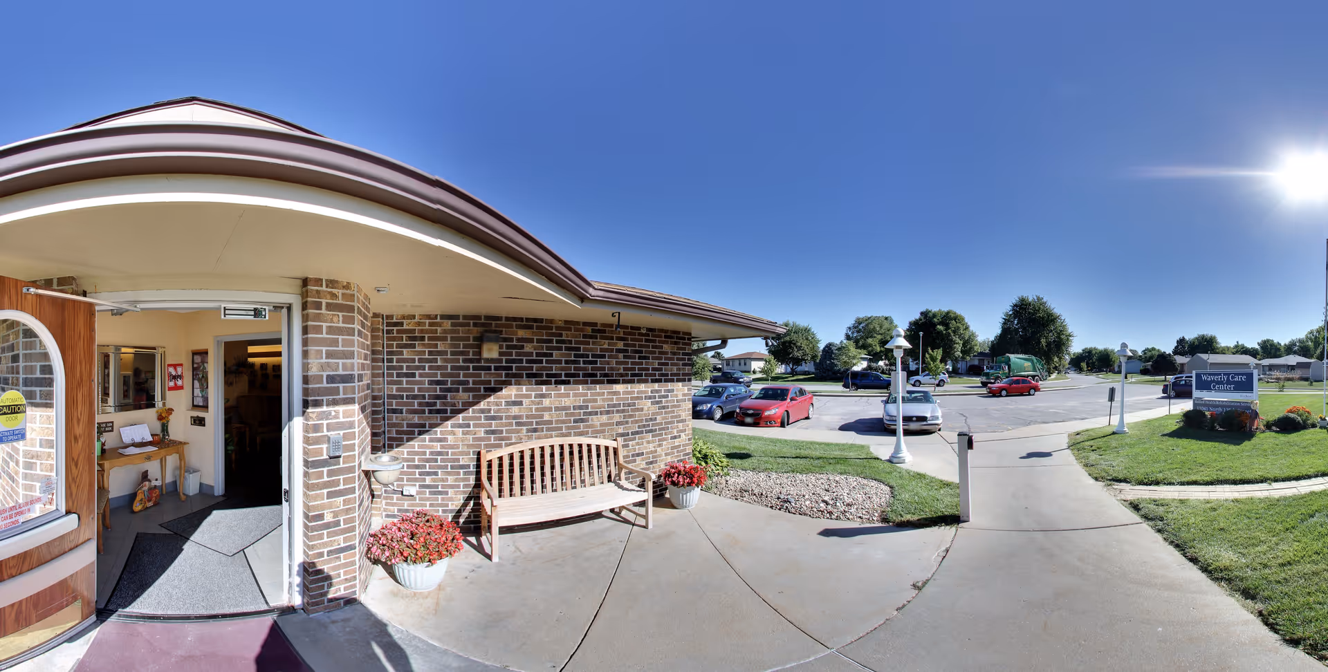Front entrance of a brick senior care facility with an open door, a wooden bench and potted flowers, and a nearby parking lot under a clear blue sky.