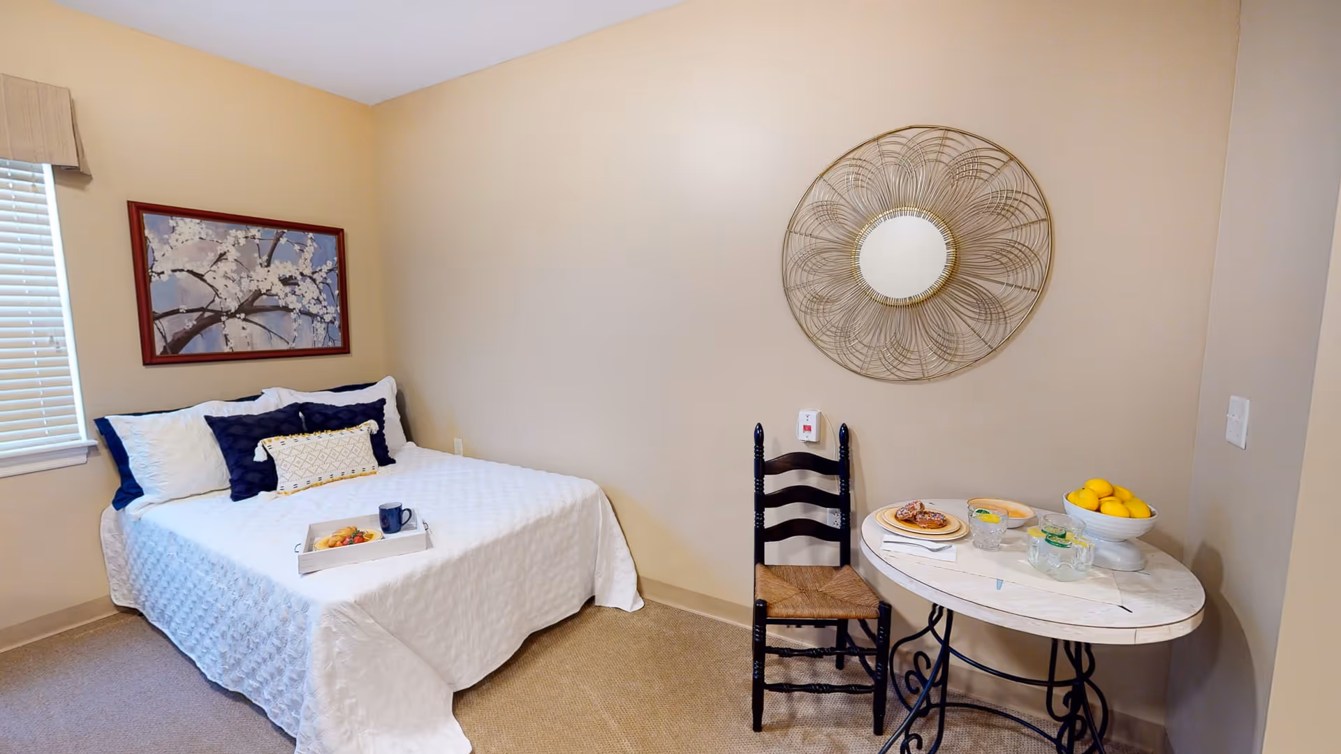 A cozy bedroom with a neatly made bed covered in a white quilt and decorated with blue and white pillows. A tray with breakfast items is placed on the bed. To the right, there is a small round table with a bowl of lemons, a plate with pastries, and two glasses of water. A wooden chair is positioned between the bed and the table. The walls are beige, adorned with a framed floral painting above the bed and a decorative round mirror above the table. A window with blinds is on the left side of the room.