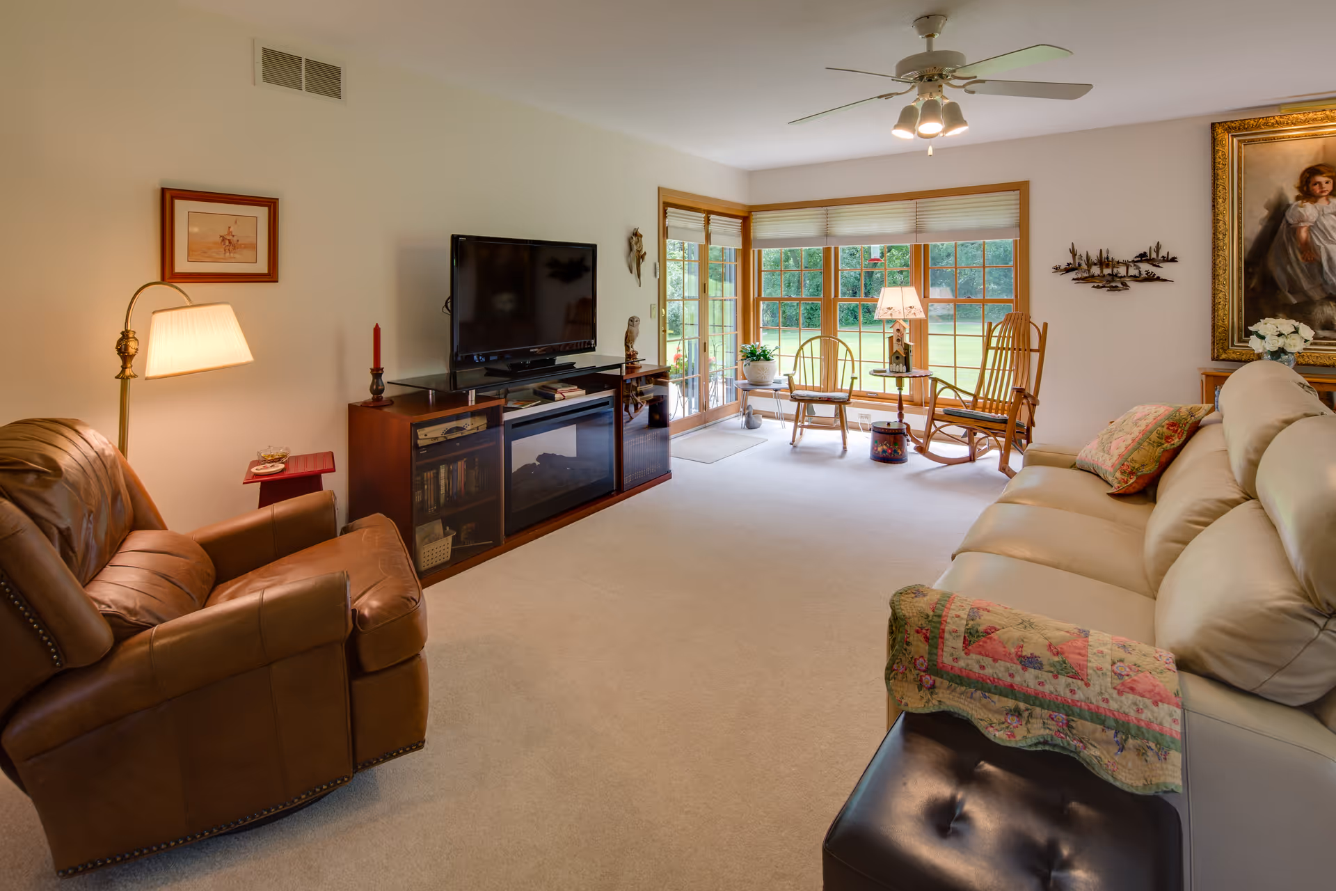 A cozy living room with a leather armchair and a cream-colored sofa adorned with a quilted throw. There is a wooden entertainment center with a flat-screen TV and decorative items. Large windows and a glass door let in natural light and offer a view of a green outdoor area. The room also features a ceiling fan, a floor lamp, wooden rocking chairs near the window, and framed artwork on the walls.