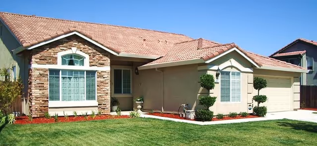 Single-story beige stucco house with a tiled roof, manicured front lawn, shrubs, and an attached two-car garage.