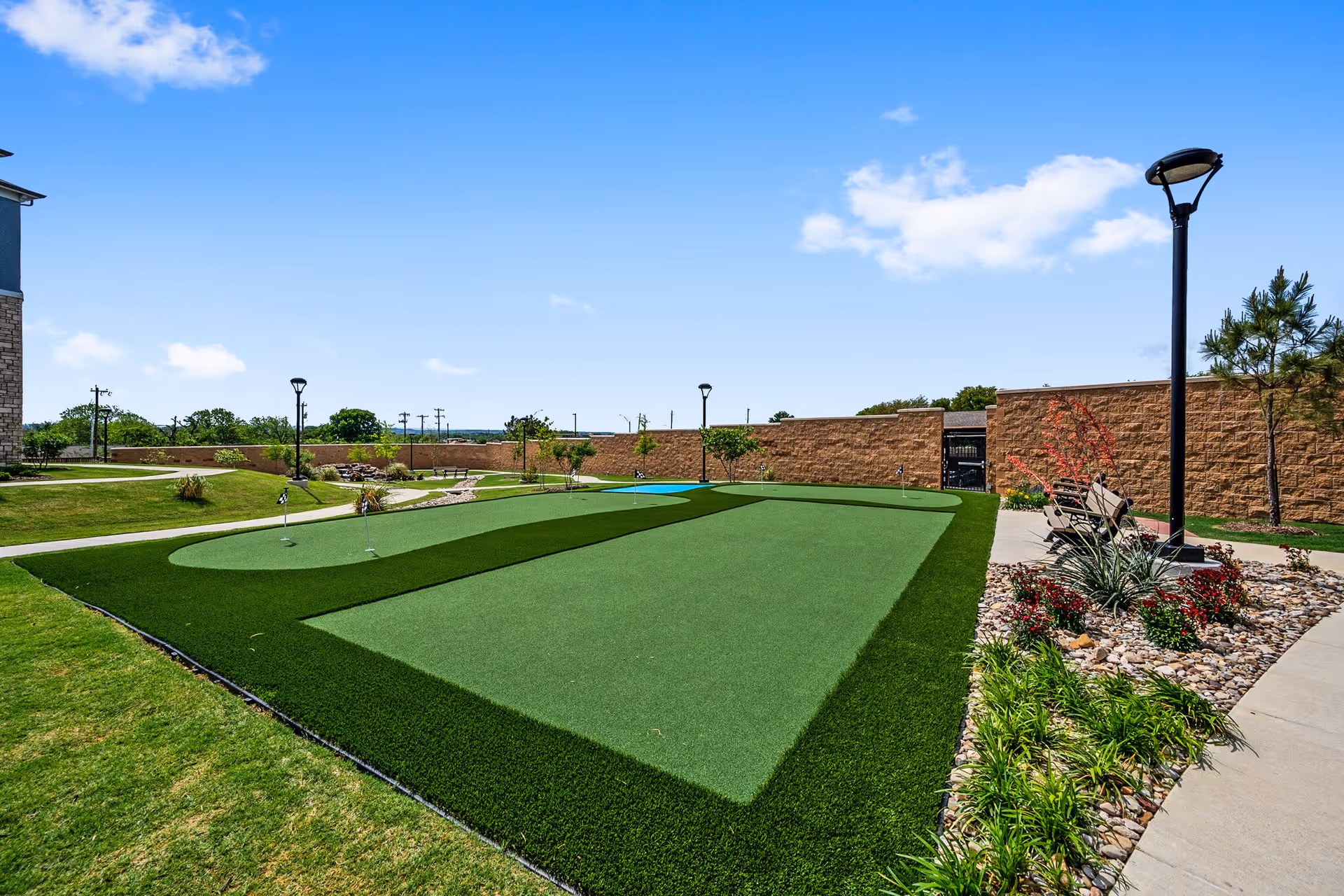 Outdoor putting green area with artificial turf, small flags marking holes, surrounded by landscaped grass, plants, benches, and lamp posts under a clear blue sky.
