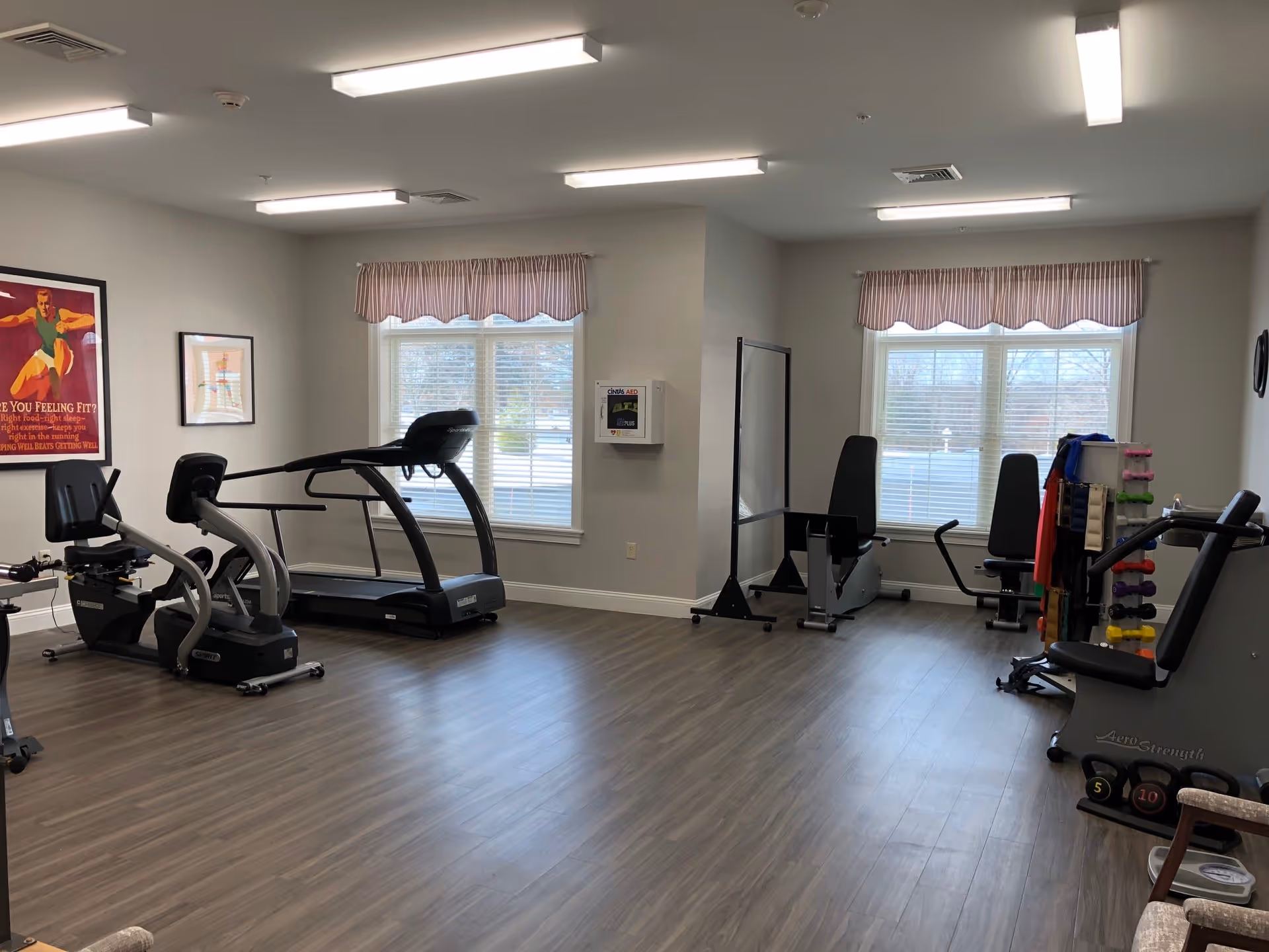 A fitness room with exercise equipment including a treadmill, stationary bike, weight machines, and dumbbells. The room has wood flooring, two windows with striped valances, fluorescent ceiling lights, and framed posters on the walls.