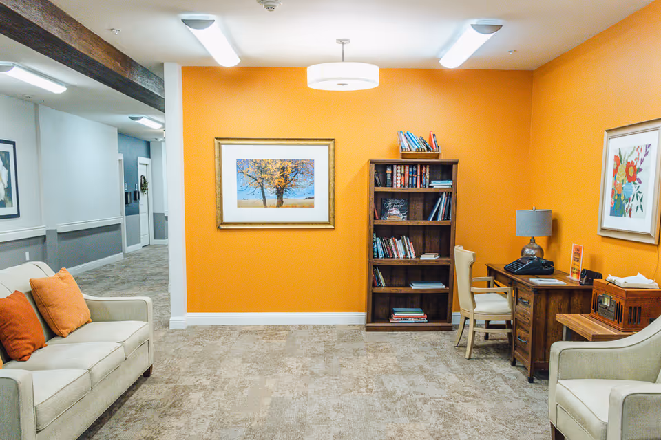A cozy interior common area with an orange accent wall featuring a framed picture of a tree. There is a wooden bookshelf filled with books, a wooden desk with a lamp, phone, and chair, and two beige armchairs with orange and red cushions. The floor is carpeted, and the adjacent hallway has gray and white walls with framed artwork.