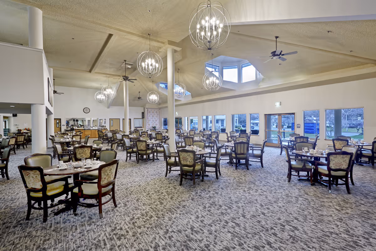 Spacious dining room with multiple round tables and upholstered chairs under decorative chandeliers and large windows.