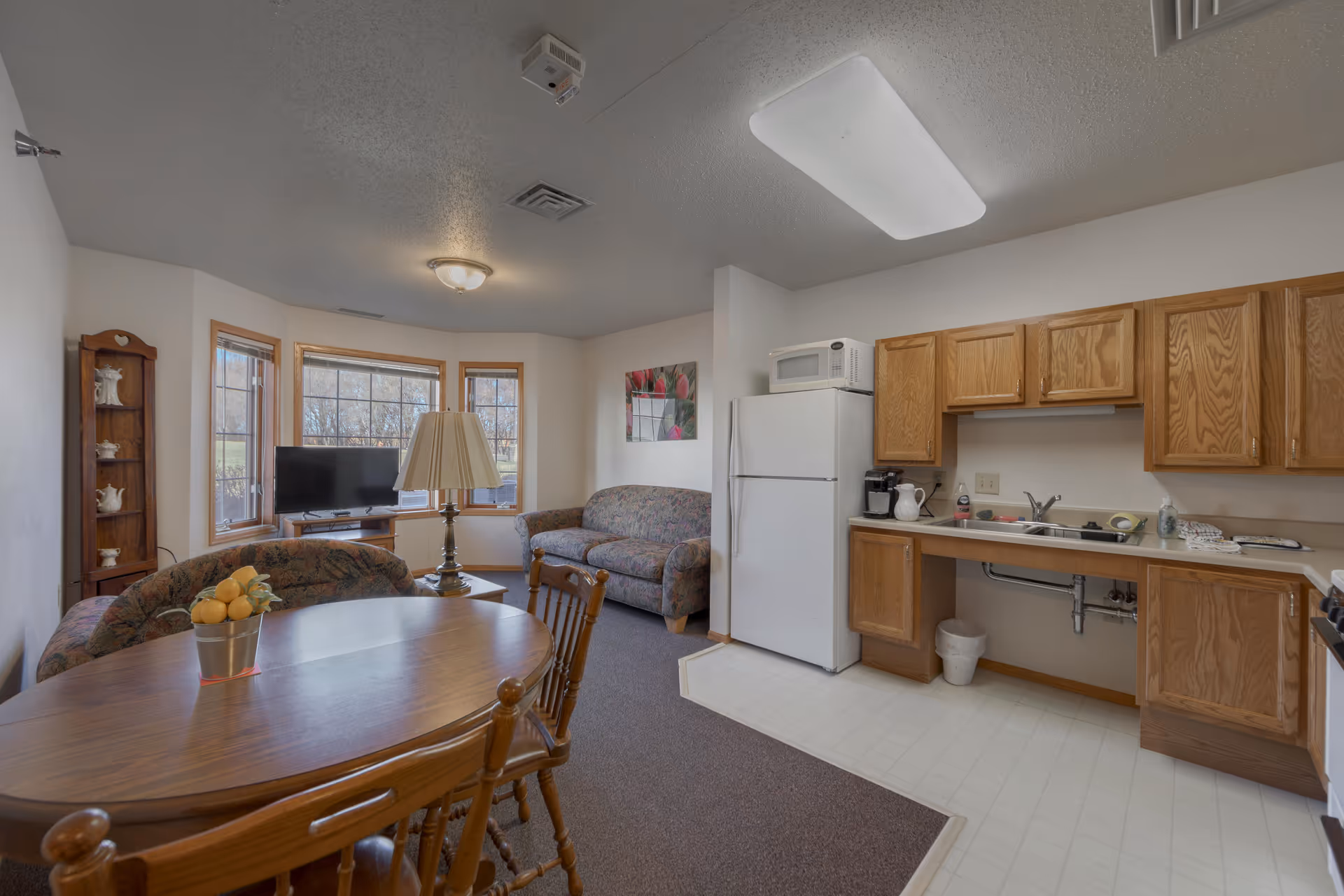 Interior view of a senior living facility room featuring a small kitchen area with wooden cabinets, a white refrigerator, microwave, and sink. Adjacent to the kitchen is a dining table with chairs and a small living area with two floral-patterned sofas, a coffee table, a floor lamp, and a TV in front of three large windows.