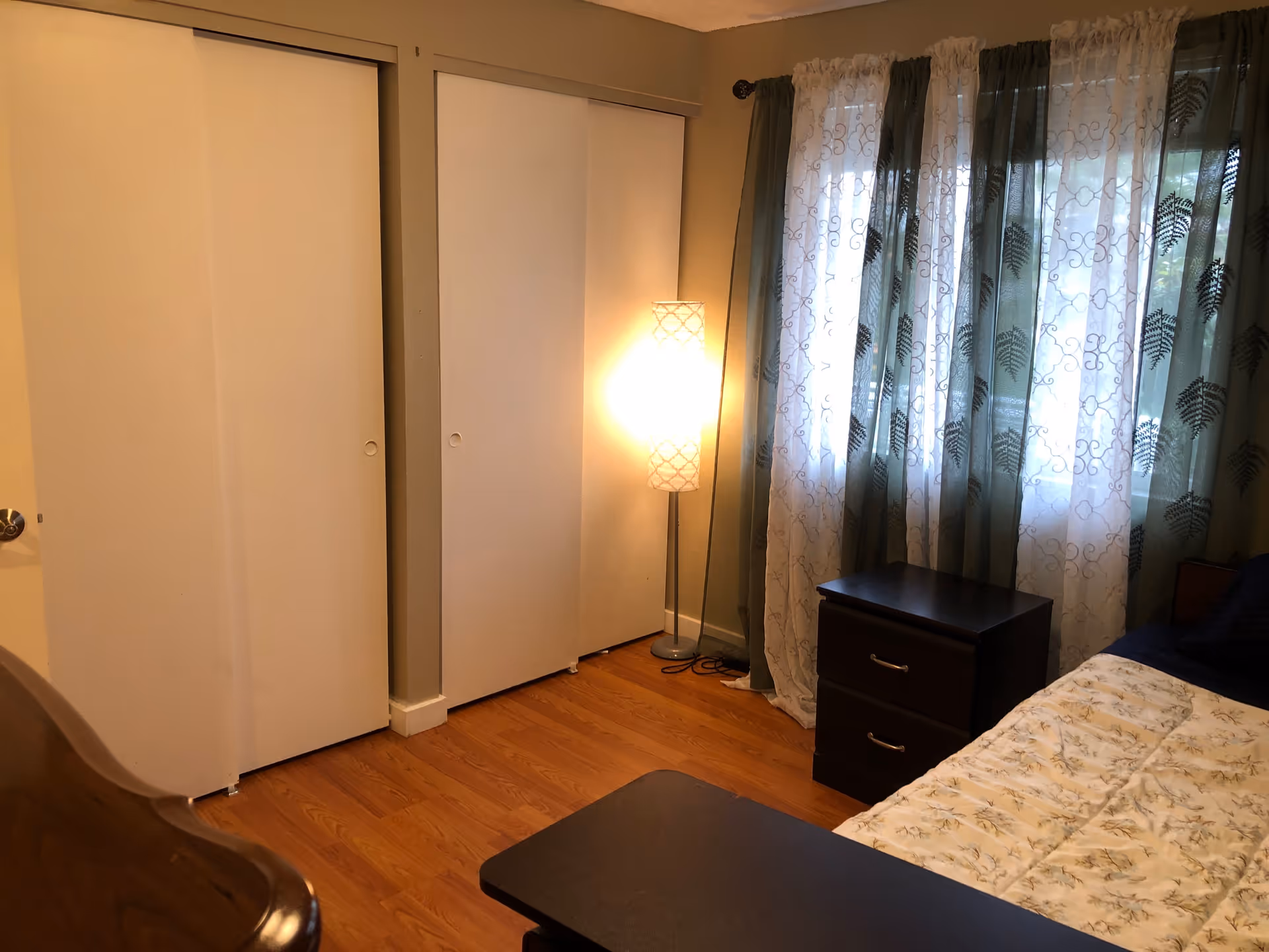 A bedroom with wooden flooring, a bed with a floral patterned comforter, a dark wooden nightstand with two drawers, a tall floor lamp emitting warm light, and a window covered with sheer white and green curtains with a leaf pattern. There are two white sliding closet doors on one wall.